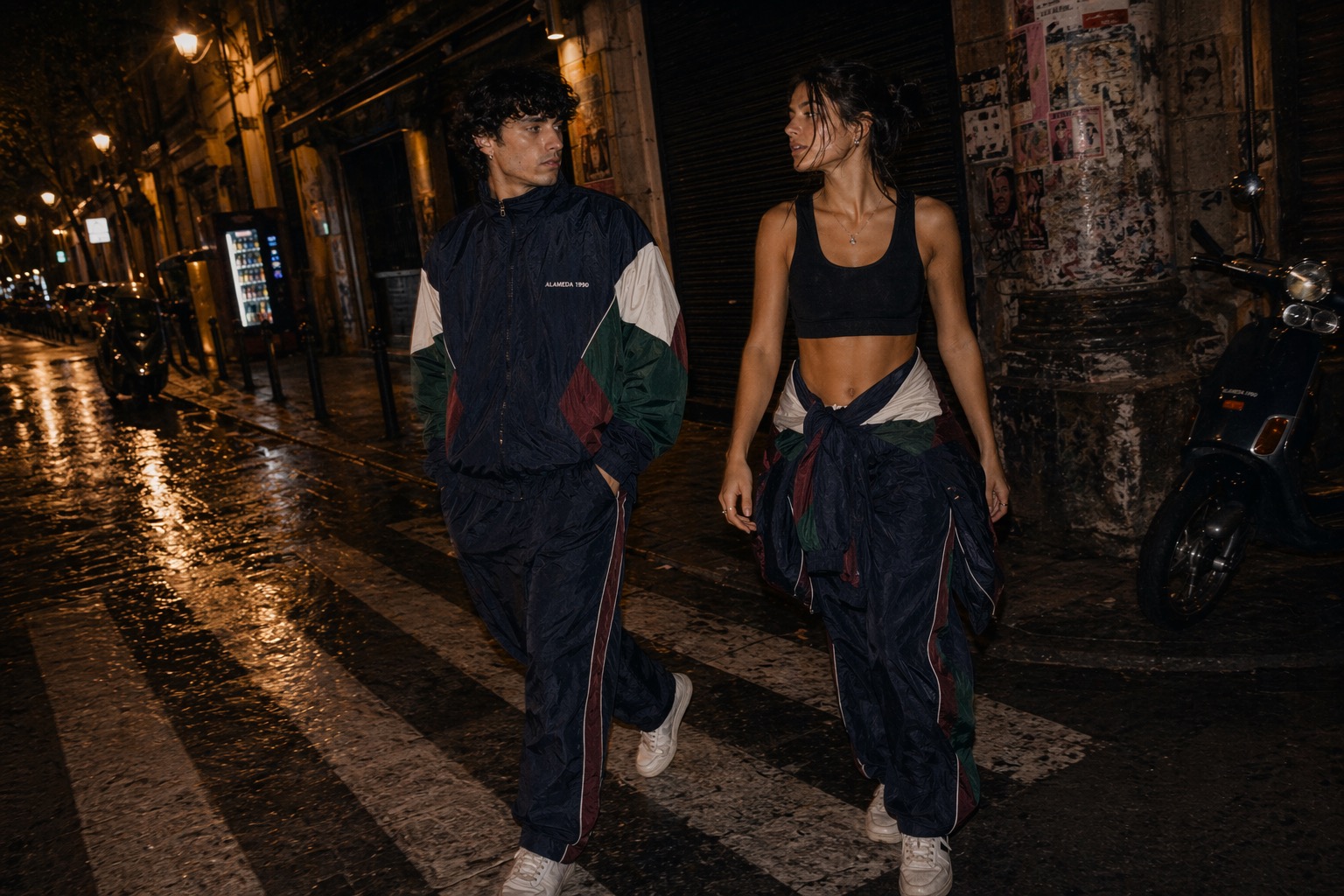 Man in a full Alameda 1990 tracksuit and woman with the tracksuit jacket tied at her waist over a black gym top crossing a wet street at night.